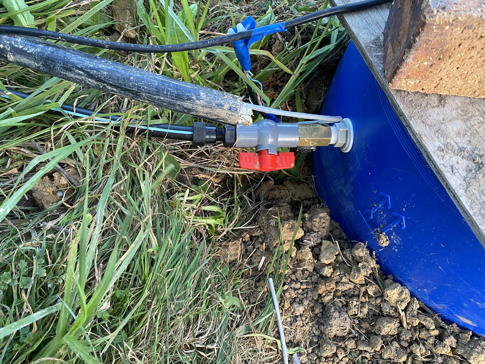 Float-valve feed line entering the blue reservoir barrel through a bulkhead fitting, with an inline brass pulse water meter and a red PVC ball valve